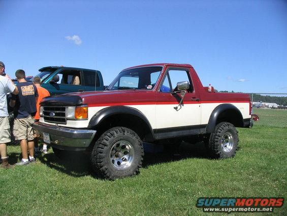 Red and white Ford Bronco with aftermarket wheels and tires parked on grass. Another truck is visible behind it