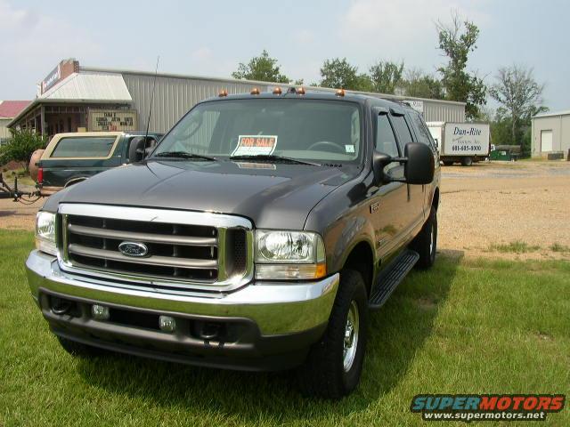 Dark gray Ford Excursion SUV parked on grass, 'FOR SALE' sign on windshield. Another SUV is visible behind it