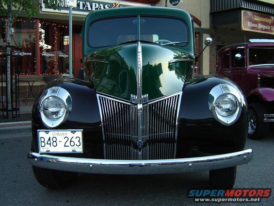 Front view of a green and black Ford truck with a chrome grille and round headlights. Another car is parked behind it