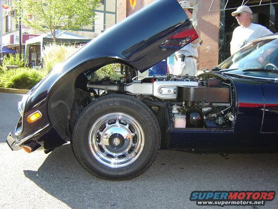 Dark blue coupe, hood open, front wheel and engine visible. Chrome trim on wheel. Two men standing nearby