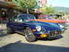 1990 Ford Thunderbird - A classic blue Jaguar E-Type coupe is parked on a street, with other vehicles and storefronts in the background.