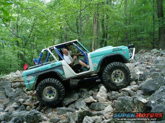 Teal and white Ford Bronco with roll cage climbing over large rocks on a wooded trail