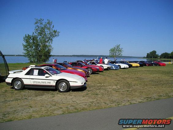 White 1984 Pontiac Fiero with "OFFICIAL RACE CAR" text on the side, parked in a line with other cars by a lake