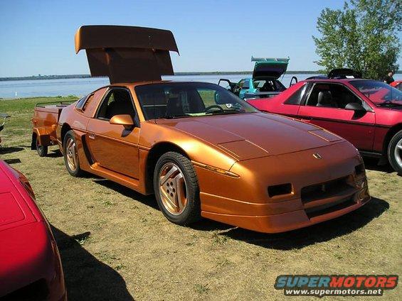 Orange 1984 Pontiac Fiero with gullwing doors open, towing a small trailer