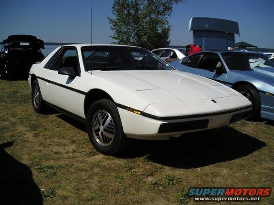 White 1984 Pontiac Fiero parked on grass, front 3/4 view. Black trim, alloy wheels, antenna. Another blue car is to the right