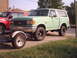 1989 Ford Bronco - A light green Ford Bronco is parked next to a red pickup truck, with a trailer hitched to the front of the Bronco.