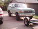 1989 Ford Bronco - A light green Ford Bronco is parked next to a red SUV, with a trailer attached to its rear.
