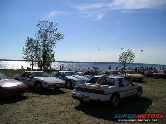Several Pontiac Fieros parked near a body of water on a sunny day. One Fiero has 'Indy Fiero' text on its rear quarter panel
