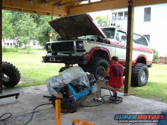 Red and white Ford Bronco with hood open, lifted with large tires, undergoing engine work