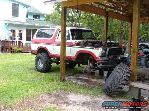 1978 Ford Bronco with hood open, undergoing repairs under a wooden shelter. Another truck is visible in the background