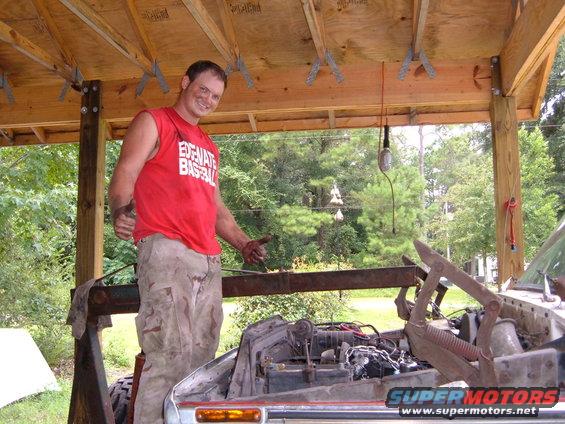 Man in red shirt and camouflage pants points to car engine under wooden structure. Supermotors.net logo visible