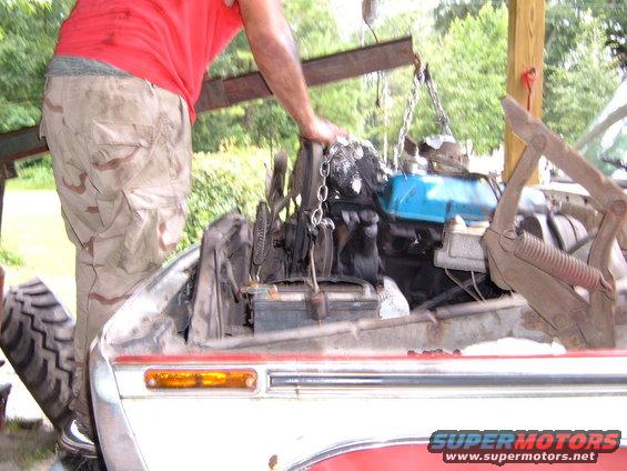 Engine being lowered into a vehicle with an engine hoist. Part of the vehicle's front fender and headlight are visible