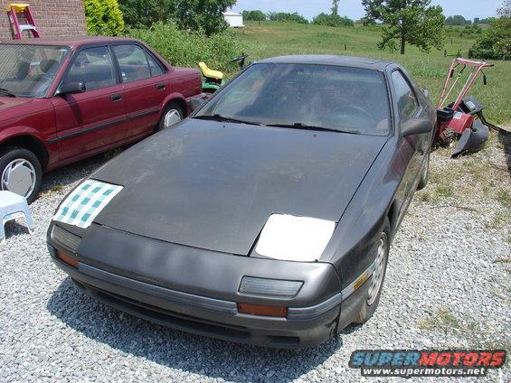 Dark gray Mazda RX-7 with pop-up headlights, one covered with a checkered cloth. Red sedan parked to the left