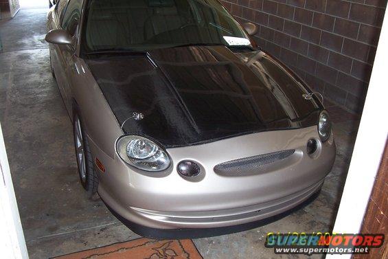 Front view of a tan car with a black carbon fiber hood and aftermarket hood pins