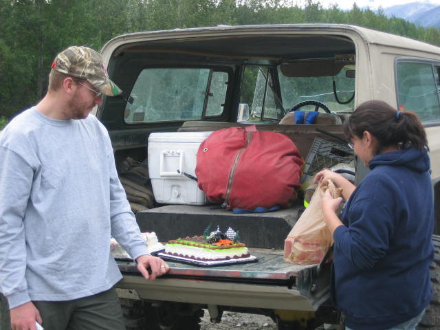 Tan SUV tailgate with a cake, cooler, and red bag; man in camouflage hat and woman in blue hoodie stand nearby