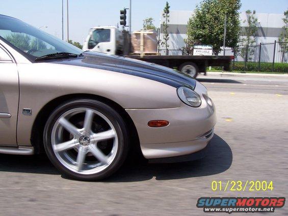 Tan car with carbon fiber hood and aftermarket wheels, front 3/4 view