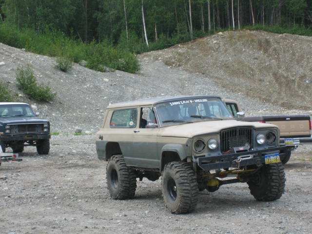 Modified Jeep Cherokee SJ with large tires and a winch, parked next to a dark blue SUV and a pickup truck