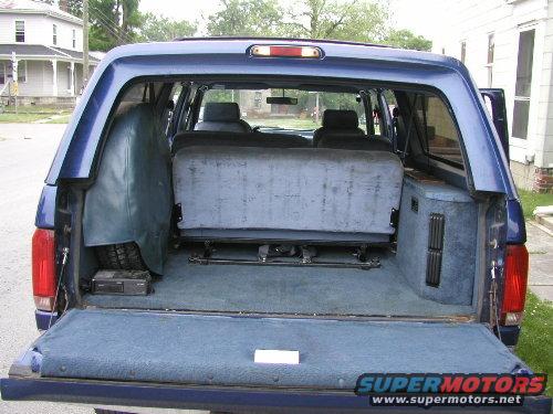 Blue van interior with folded rear seats and spare tire. Supermotors.net logo visible