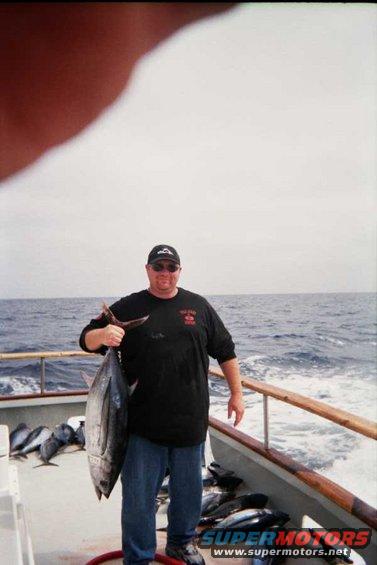 Man in black shirt and blue jeans holding a large tuna on a boat deck with other fish