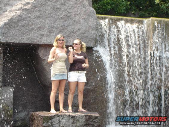 Two women stand on rocks near a waterfall. Water cascades down a stone wall