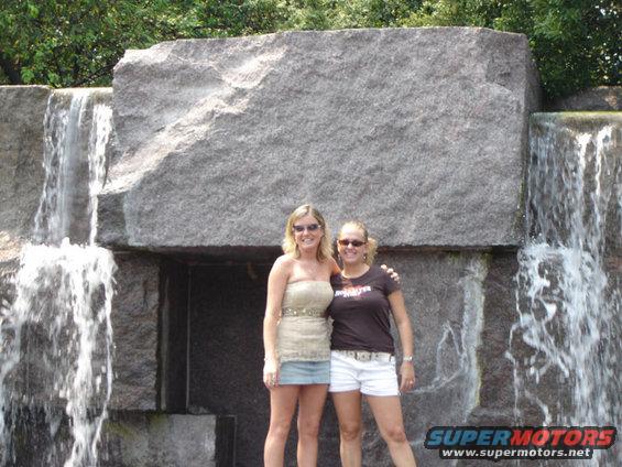 Two women stand in front of a large rock with waterfalls on either side. One wears a strapless top and denim skirt, the other a t-shirt and white shorts