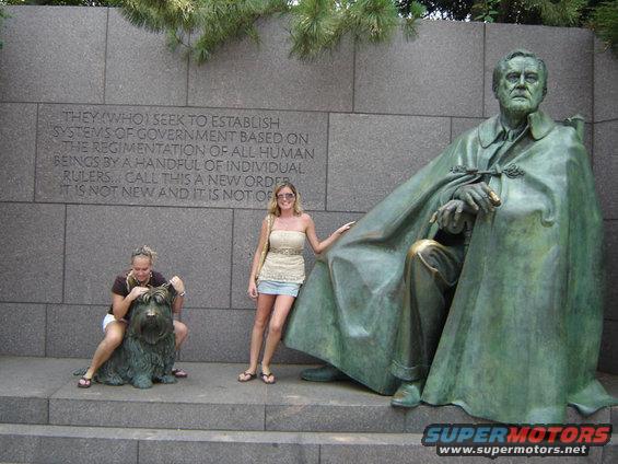 Bronze statue of a seated man in a cape, with two women and a dog statue nearby