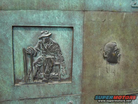 bronze relief sculpture showing a seated man in a hat holding a tool, and a profile of a head
