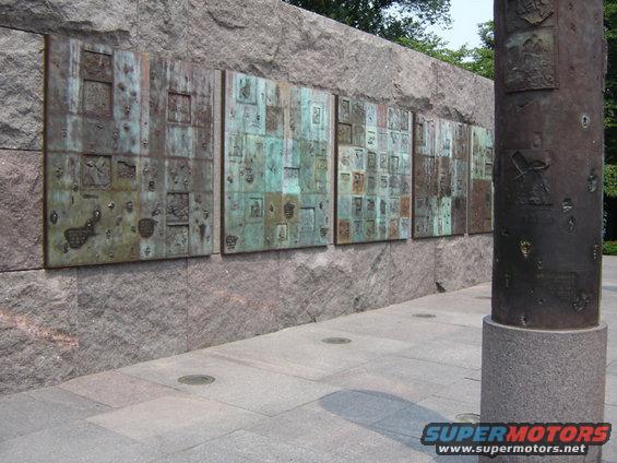 bronze relief panels on a stone wall, with a cylindrical monument to the right