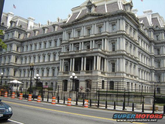 Large ornate building with columns and many windows, fronted by a fence and street with traffic barrels