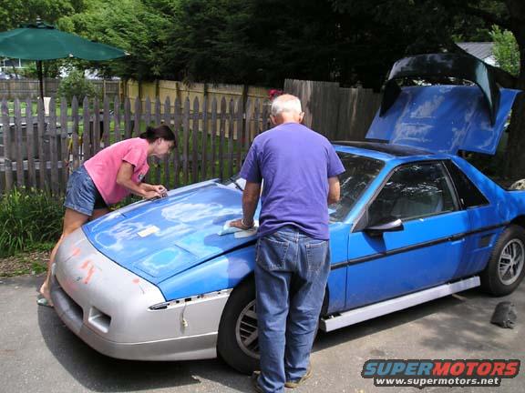 Two people sanding and prepping a blue Pontiac Fiero with raised engine hatch