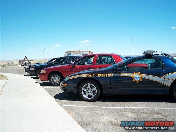 Three police cars parked side-by-side: a black Ford Crown Victoria, a red sedan, and a dark blue Chevrolet Camaro with 'STATE TROOPER' text
