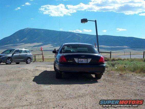 Black 2001 Ford Crown Victoria from rear, license plate ADAM-12, parked on dirt lot with another car and mountains in background