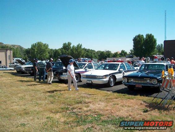 Several cars parked in a line on a grassy field under a clear blue sky, with people milling around