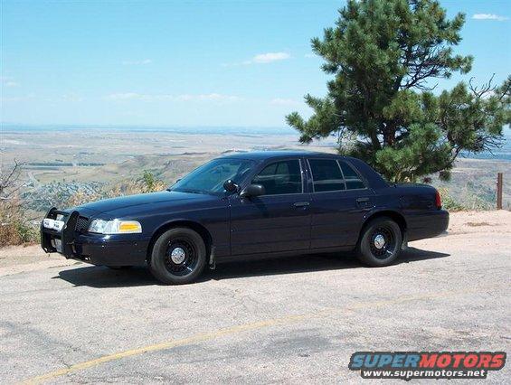Dark sedan with a push bumper parked on a hill overlooking a valley. It looks like a 2001 Ford Crown Victoria