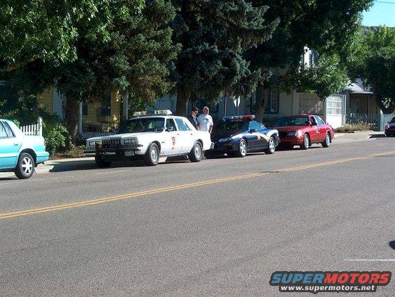Three police cars parked on a street, a white car in front, a dark blue car in the middle, and a red car in the rear