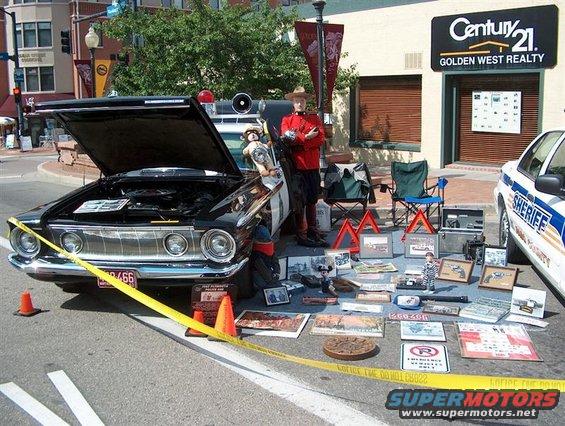Black vintage police car with hood open, display of items on street, and a sheriff's car parked nearby