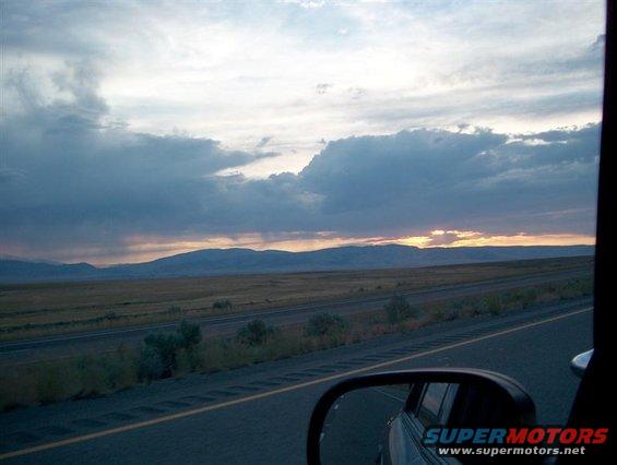 Side mirror reflecting a highway and distant mountains under a cloudy sky at sunset