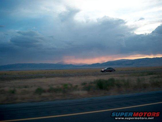 Sedan driving on a highway with mountains in the background under a cloudy sunset sky. Supermotors.net logo visible