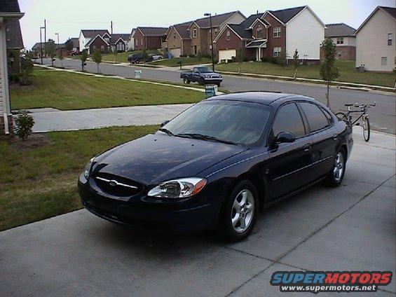 Dark blue sedan parked in driveway with houses in background. Bicycle on rack behind it