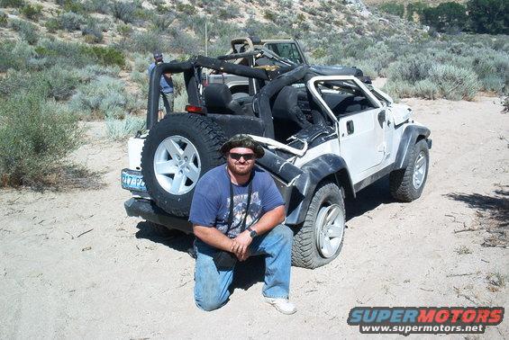 Man kneeling in front of a white Jeep Wrangler with a spare tire on the back. Another Jeep is behind it