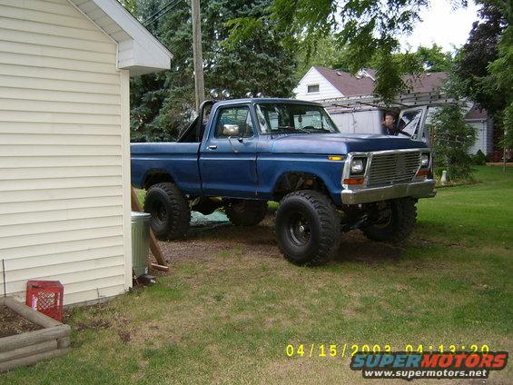Blue lifted pickup truck parked next to a house. Someone is in the driver's seat with the door open