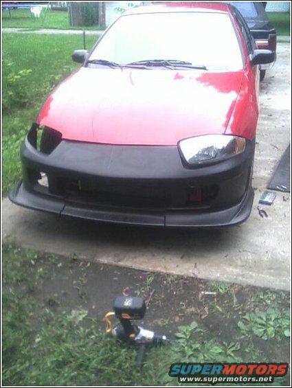 Red car with a large black aftermarket bumper and front lip, parked on a concrete driveway with grass