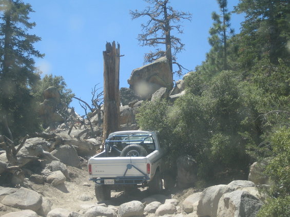 White Ford Bronco with spare tire in bed driving on a rocky, wooded trail. License plate visible