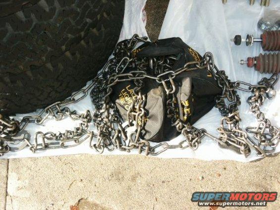 Tire chains and shock absorbers on a concrete surface. A black bag with yellow lettering is visible