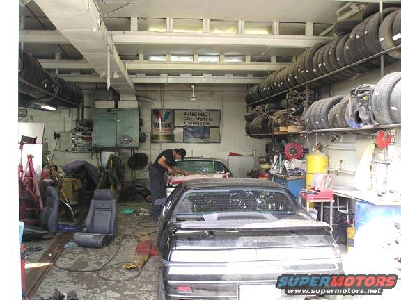 Black Pontiac Fiero in a garage with tires stacked on shelves and a mechanic working on another car