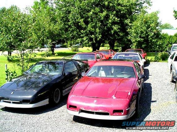 Black Pontiac Fiero with white front bumper, parked on gravel