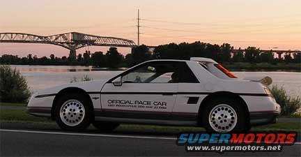 White 1984 Pontiac Fiero with 'Official Pace Car' decals parked by a river with a bridge in the background at sunset