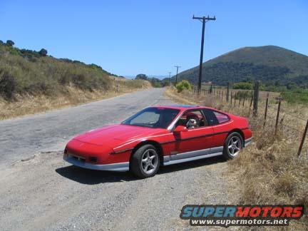 Red 1986 Pontiac Fiero parked on a dirt road with a grassy hill and a mountain in the background