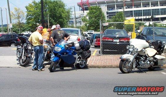 Three motorcycles parked on asphalt, with people standing around two of them. A white motorcycle is to the right