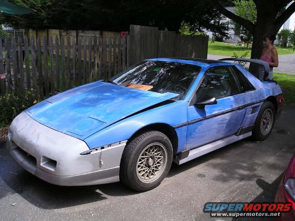 Blue Pontiac Fiero with primer-colored front bumper and hood, undergoing repairs